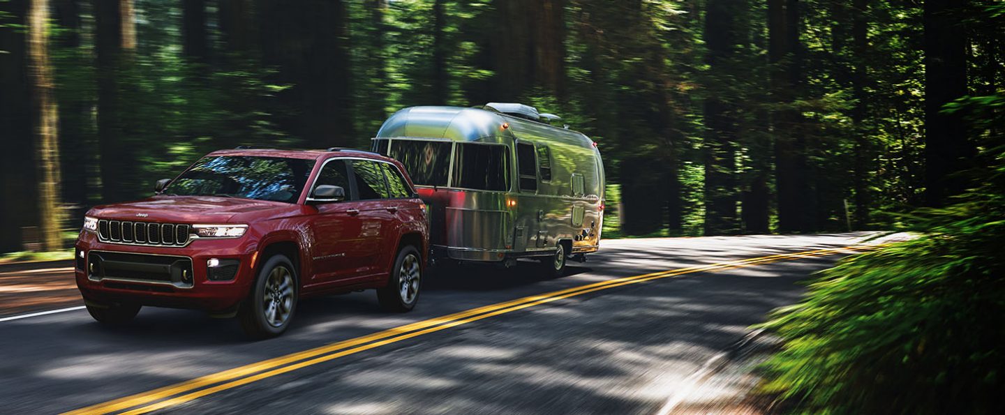 A 2022 Jeep Grand Cherokee Overland towing a travel trailer as it's driven down a forest road.