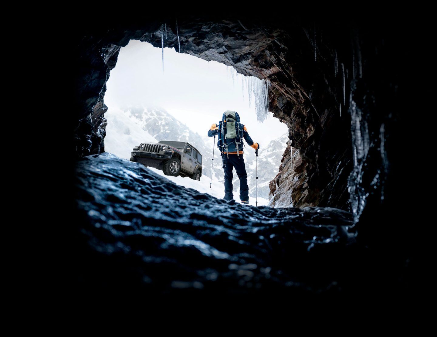 A bundled-up hiker with walking poles approaches a 2022 Jeep Wrangler Rubicon parked on a snowy mountainside beside an ice cave.
